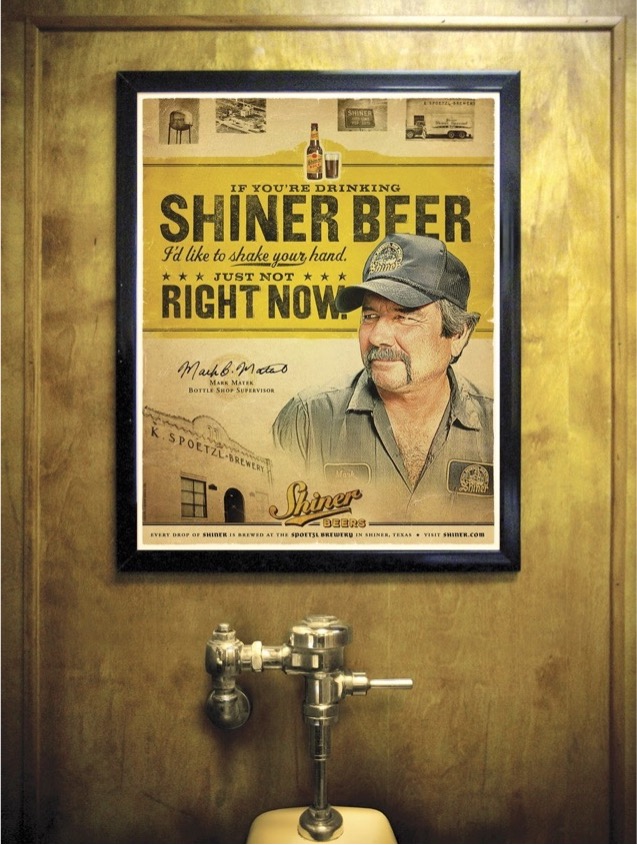 A framed Shiner Beer poster above a urinal reads ‘If you’re drinking Shiner Beer, I’d like to shake your hand—just not right now,’ playing on the setting.