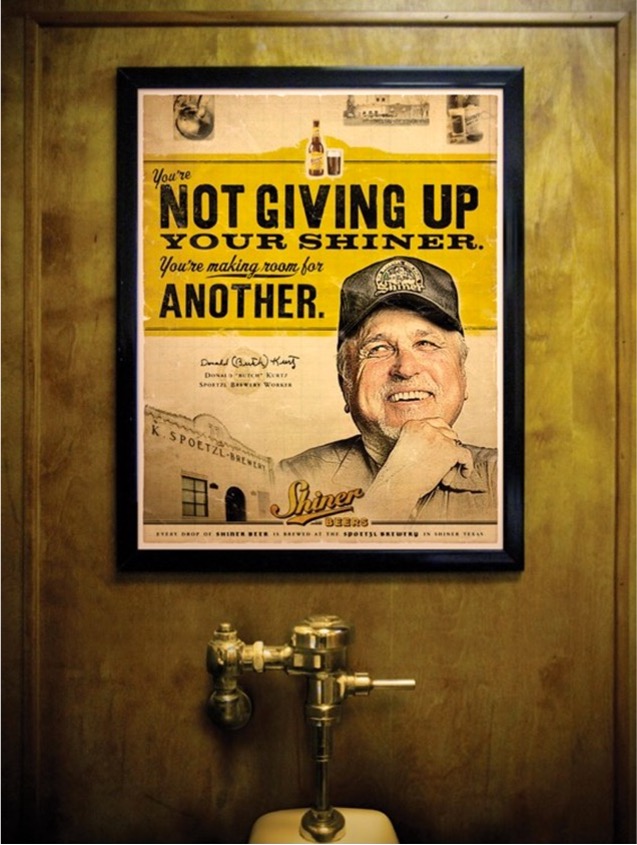 A Shiner Beer poster above a urinal reads ‘You’re not giving up your Shiner. You’re making room for another,’ playing on the bathroom setting.